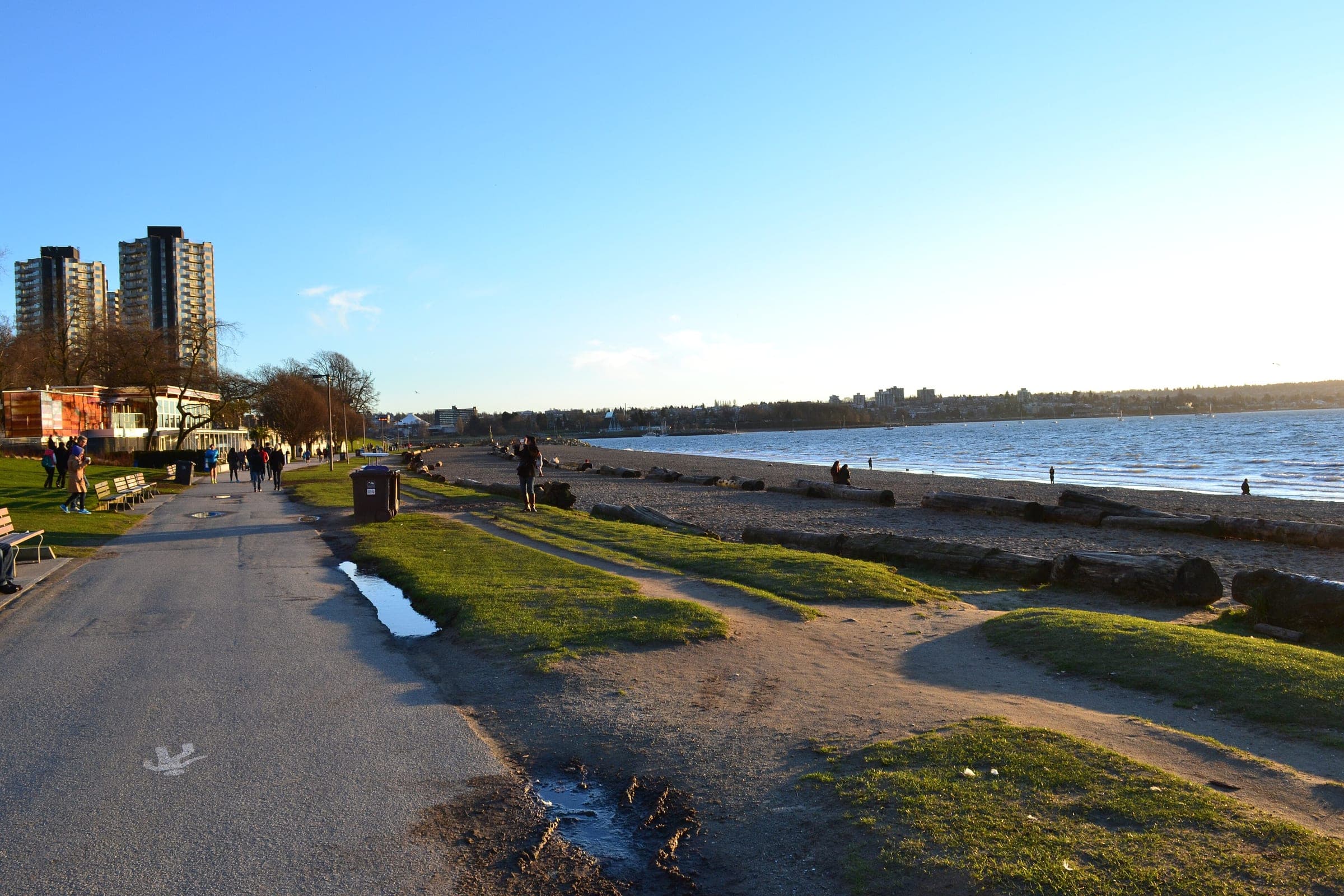 Walkers follow the seawall path past the logs on English Bay Beach in low February sun.