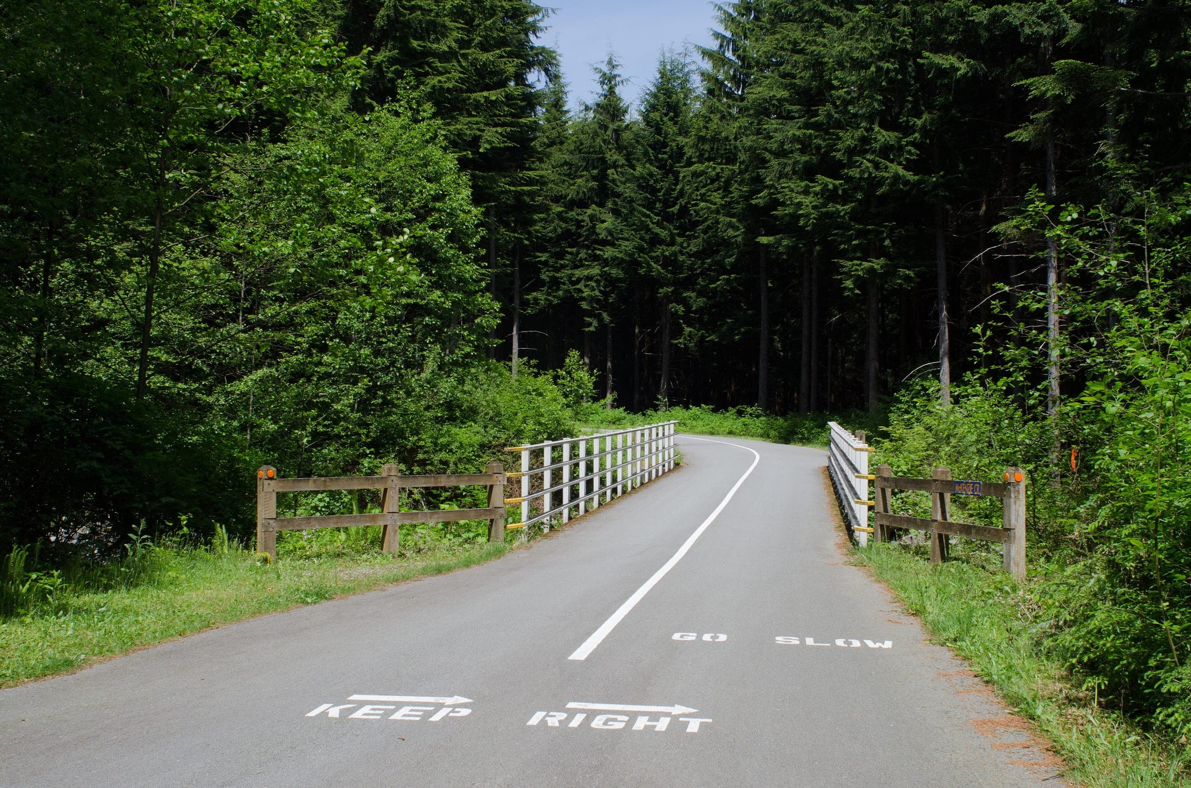 The paved Seymour Valley Trailway crosses a fenced bridge into tall forest in the Lower Seymour Conservation Reserve.