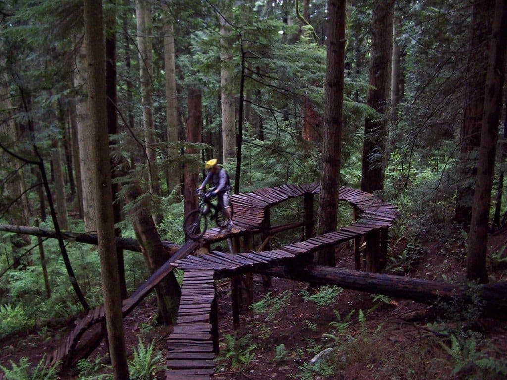 A rider rolls a raised wooden switchback bridge through the cedar forest on Mount Fromme.