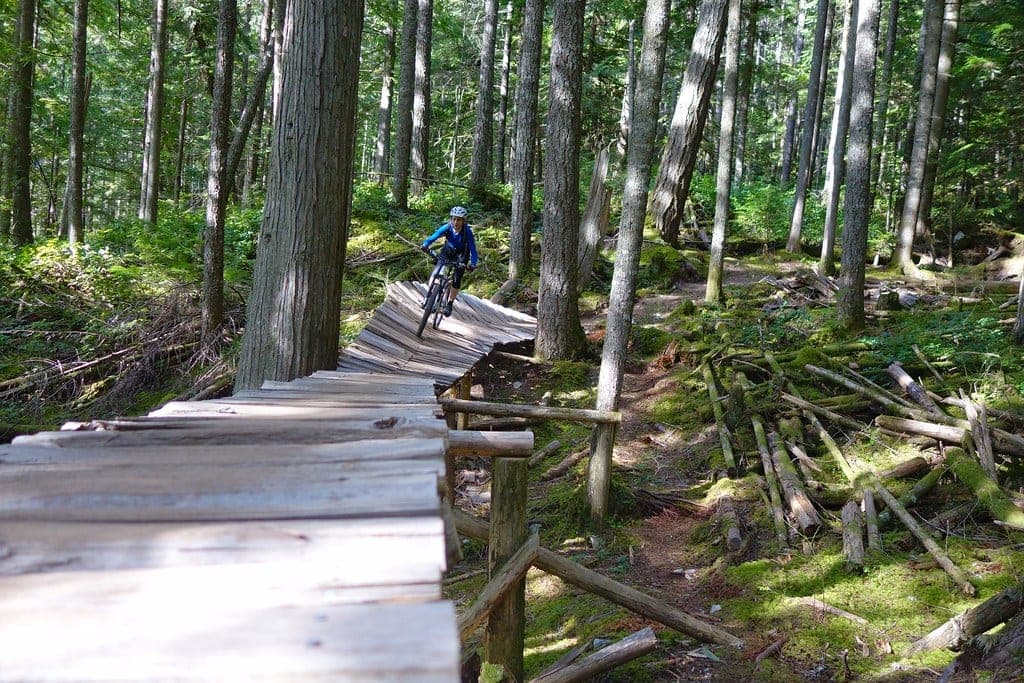 A rider crosses a long wooden bridge through sunny second-growth forest on a SORCA trail in Squamish.