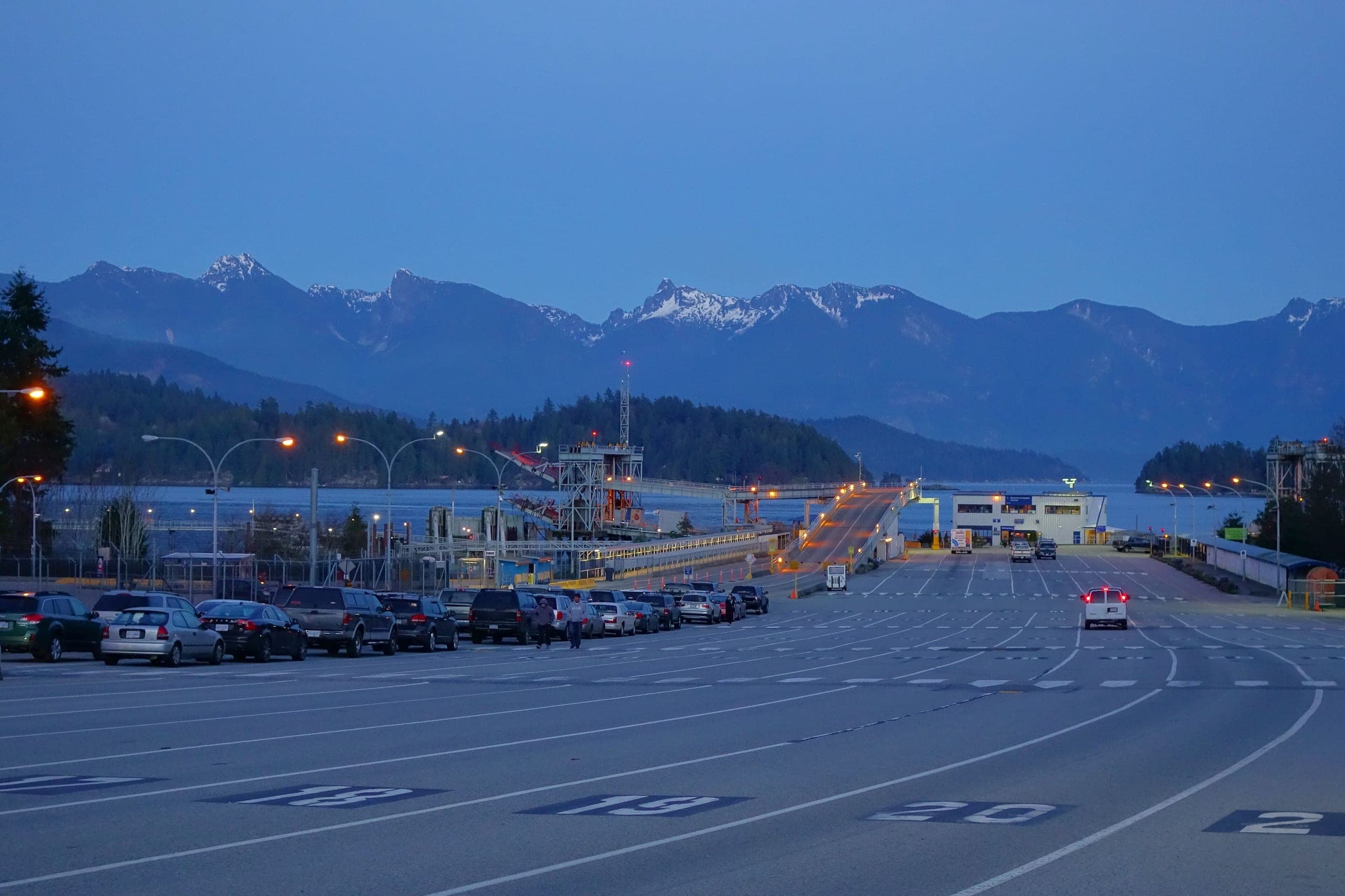Cars queue at the BC Ferries Langdale terminal at dusk with snow on the ridges across Howe Sound.