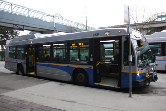 A blue and white TransLink hybrid bus waits at a Metro Vancouver bus loop beside the SkyTrain guideway.
