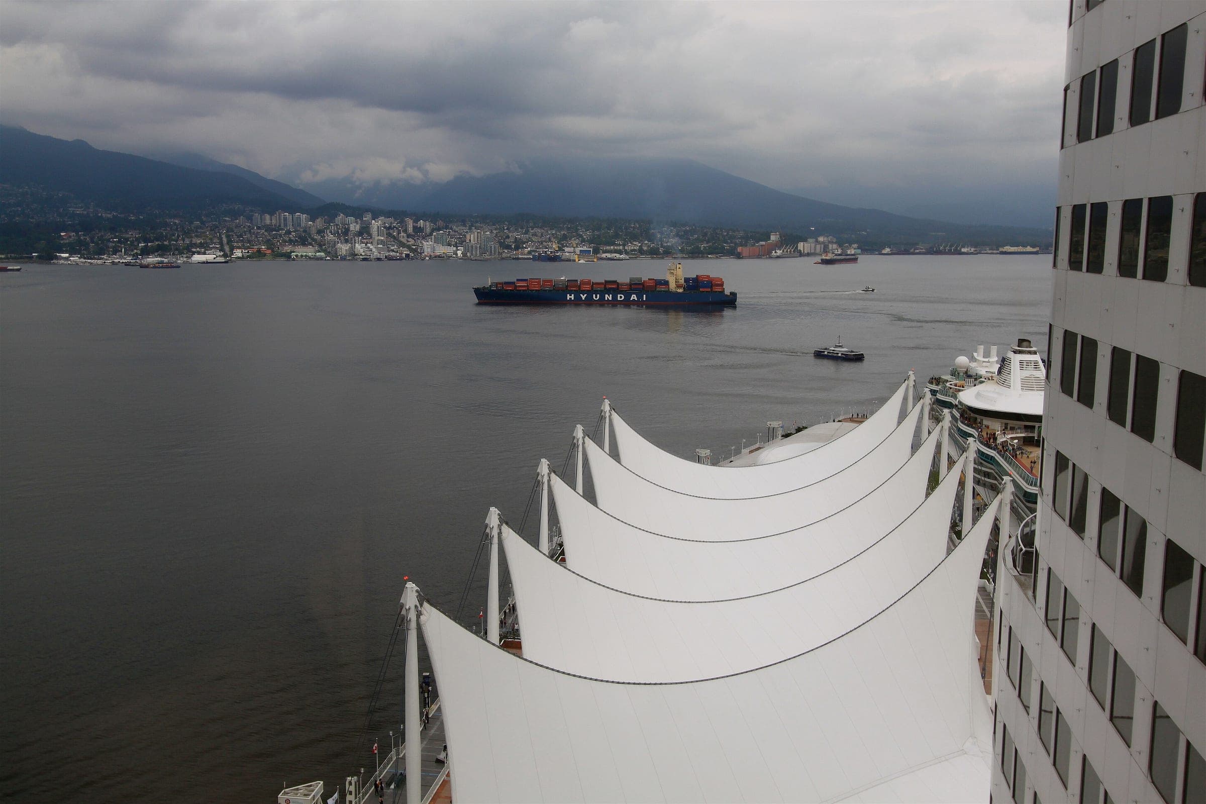 The sail roof of Canada Place overlooks Burrard Inlet and a ship at anchor under heavy cloud.