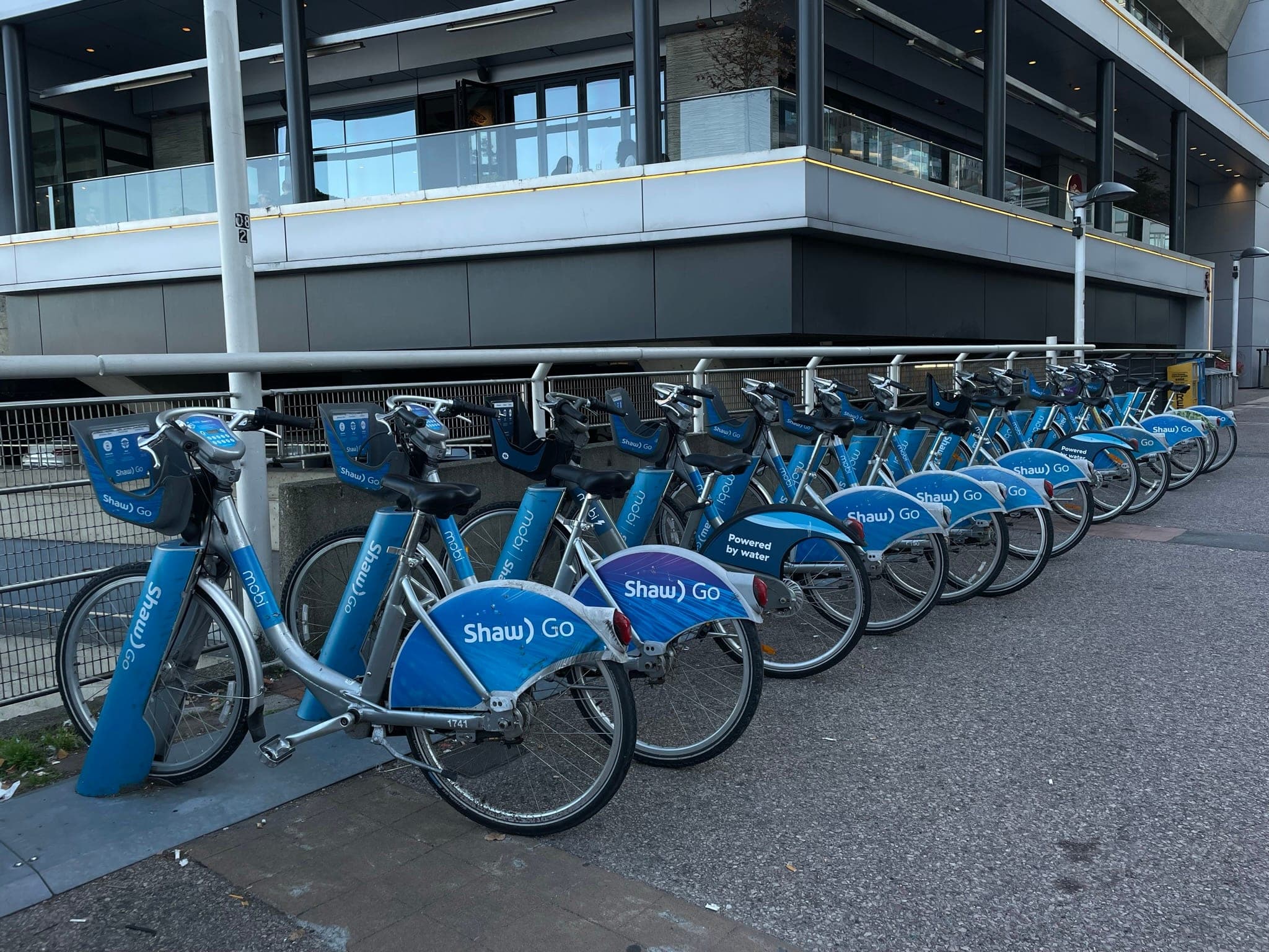 Blue Mobi share bikes line a docking station beside a waterfront building in Vancouver.